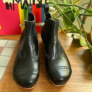 May include: A pair of black leather ankle boots with decorative stitching and wooden soles. The boots have a pull tab at the back and are set on a wooden surface. The boots are a classic style.