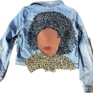 Op de afbeelding: Een blauwe denimjas met een geschilderd ontwerp op de rug. Het ontwerp toont een vrouw met een bruine huid, zwart krullend haar en een shirt met luipaardprint.