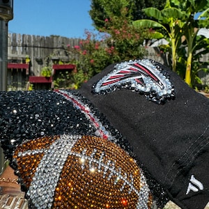 May include: A black baseball cap with a rhinestone football design. The football is brown and white with a red and white Atlanta Falcons logo on the front.