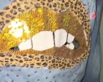 Op de afbeelding: Lichtblauwe spijkerjas met een groot pailletten lippenontwerp. De lippen zijn goud en wit, met een luipaardprint rand. De jas is versierd met kleine, kleurrijke strass steentjes en roze bloemenaccenten.