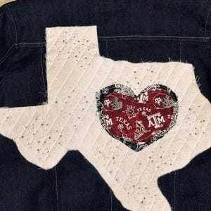 May include: Dark blue denim jacket with a white quilted Texas-shaped patch. The patch is embellished with rhinestones and features a heart-shaped design with "Texas" and "ATM" text in maroon and black.