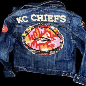 Puede incluir: Chaqueta vaquera azul con las palabras "KC CHIEFS" en la parte superior de la espalda. La chaqueta presenta un gran diseño de labios con lentejuelas con el logotipo de los Chiefs. Parches del logotipo de los Chiefs en la parte delantera y la manga.
