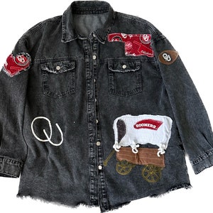 May include: A black denim jacket with patches and embroidery. The patches feature the words "Oklahoma" and "Sooners" in red and white. The embroidery includes a white "Q" and a wagon wheel design.