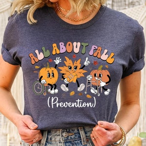 May include: A navy blue t-shirt with the text "ALL ABOUT FALL" in a retro font. The shirt features cartoon pumpkins and leaves with the word "(Prevention)" below the graphic. The shirt is worn by a person.