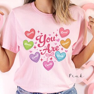 May include: A light pink short-sleeved t-shirt featuring a graphic of six colorful heart-shaped candies surrounding the text You Are. The hearts are red, green, purple, pink, orange, and blue, each inscribed with words including Strong, Brave, Enough, Capable, Worth, Unique, and Loved.