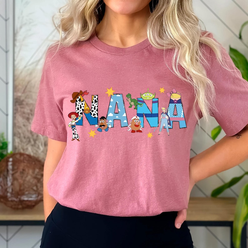 May include: A dusty rose t-shirt with the word "NANA" in a colorful, cartoon-style design. The letters are decorated with characters from a popular animated movie. Yellow stars are scattered around the letters.