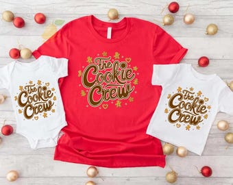 May include: A bright red short-sleeved t-shirt, a white short-sleeved t-shirt, and a white short-sleeved bodysuit. Each garment features the brown text The Cookie Crew surrounded by small gold gingerbread man and star graphics printed on the front.