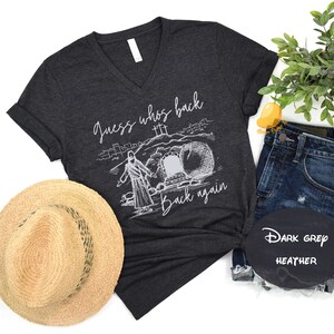 May include: Dark grey heather v-neck t-shirt with a white graphic of a man walking out of a tomb with the text "Guess who's back... back again".
