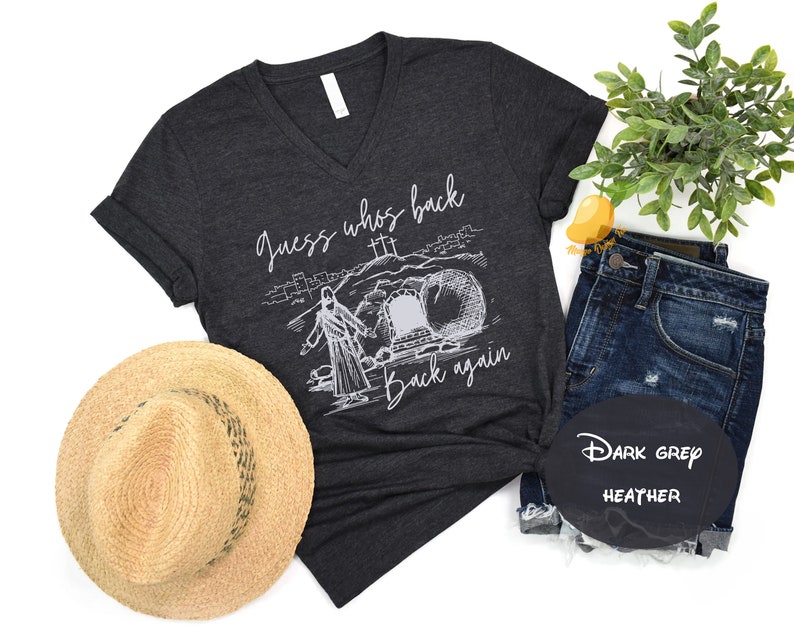 May include: Dark grey heather v-neck t-shirt with a white graphic of a man walking out of a tomb with the text "Guess who's back... back again".