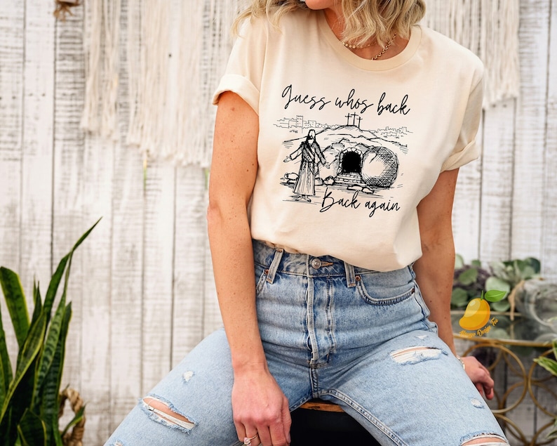 May include: A beige t-shirt with a black graphic of Jesus walking out of a tomb with the text "Guess who's back... back again".