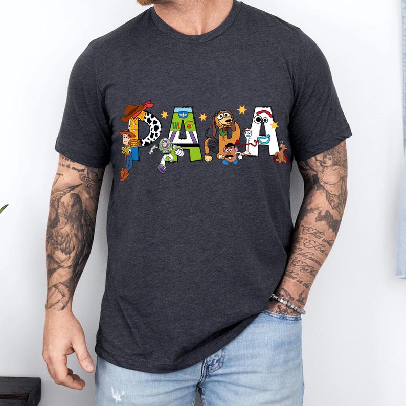 May include: Dark gray t-shirt with the word "DADA" in colorful letters, each decorated with characters from a popular animated movie. The letter "D" features a cowboy, the "A" has a space ranger, and the other letters have a dog, a potato head, and a snowman.