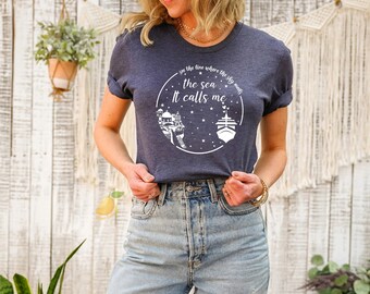 May include: A heather navy blue short-sleeve t-shirt featuring a white circular graphic with stars, a ship, and a building. The text reads, "see the line where the sky meets the sea It calls me" in a white script and sans-serif font combination.