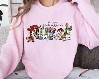 May include: A light pink crewneck sweatshirt featuring the text "pediatric NURSE" in black script and block lettering. The letters are filled with colorful cartoon character graphics, including a cowgirl hat, space ranger, and various toys.