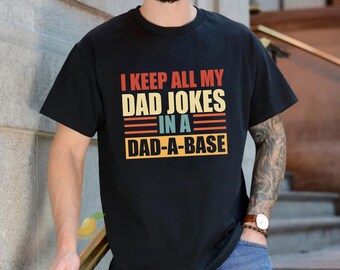 May include: A black short-sleeved t-shirt featuring a graphic print with the text I KEEP ALL MY DAD JOKES IN A DAD-A-BASE. The text is displayed in a retro-style font with alternating lines of orange, yellow, and teal against the black fabric.