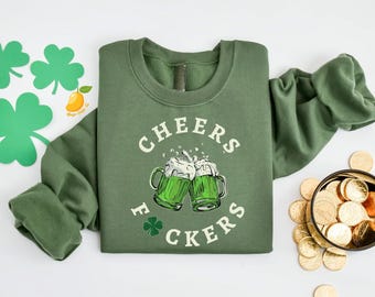 May include: An olive green crewneck sweatshirt featuring a graphic of two clinking beer mugs filled with green liquid and white foam. The text "CHEERS F*CKERS" is printed in a circular arc around the mugs, with a small green shamrock icon.