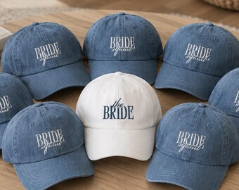 May include: A collection of baseball caps featuring one white cap with the text the BRIDE in dark blue and several blue denim caps with the text BRIDE squad embroidered in white. Each cap has a curved brim and a structured crown design.