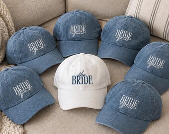 May include: A collection of six blue denim baseball caps and one white baseball cap. Each blue cap features the white embroidered text "BRIDE squad" in a script font. The white cap features the dark blue embroidered text "the BRIDE" in a serif font.