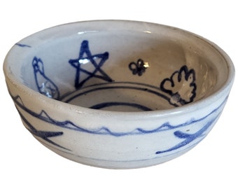 May include: A small white ceramic bowl with a blue and white design. The design features a star, a flower, a bird, and a leaf. The bowl has a wavy rim.