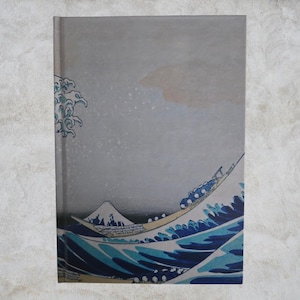 Puede incluir: Un cuaderno con una impresión de "La Gran Ola de Kanagawa" de Hokusai. La portada representa una gran ola oceánica con un barco y el monte Fuji al fondo. La paleta de colores incluye tonos de azul, blanco y gris.
