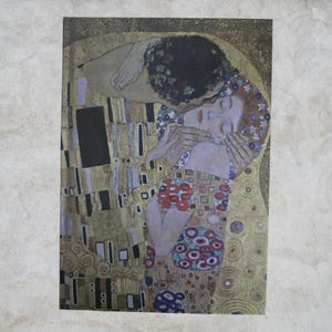 Puede incluir: Una impresión de "El Beso" de Gustav Klimt muestra a una pareja abrazándose, con el rostro del hombre oculto. La obra de arte es rica en tonos dorados y tierra, con patrones y detalles intrincados. El vestido de la mujer tiene patrones florales rojos y azules.