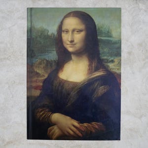 Puede incluir: Un cuaderno con la pintura de la Mona Lisa de Leonardo da Vinci en la portada. La imagen muestra un retrato de una mujer con una sonrisa sutil, sobre un fondo de paisaje. La paleta de colores incluye verdes, azules y marrones apagados, creando una estética clásica.