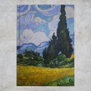 Puede incluir: Un cuaderno con una impresión de "Campo de trigo con cipreses" de Vincent van Gogh. La portada muestra un paisaje vibrante con un campo amarillo, árboles verdes, montañas azules y un cielo arremolinado. La obra es una reproducción.