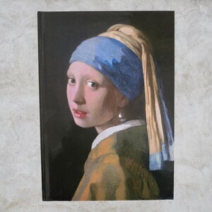 Puede incluir: Un cuaderno con una impresión de "La joven de la perla" de Johannes Vermeer. La obra de arte representa a una joven con un pañuelo azul y amarillo, un pendiente de perla y una chaqueta color mostaza. El fondo es negro.