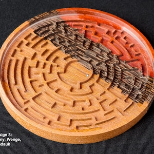 May include: A circular wooden maze with a red, brown, and black wood design. The maze is made of three different types of wood: mahogany, wenge, and padauk. The text "Design 3: Mahogany, Wenge, Padauk" is below the maze.
