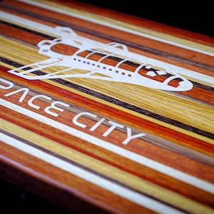 以下が含まれることがあります： スペースシャトルのデザインと「SPACE CITY」という文字が刻まれた木製のまな板。複数の種類の木材で作られており、カラフルなストライプ模様になっています。