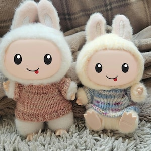 May include: Two plush bunny dolls with smiling faces and small pink tongues. They wear knitted sweaters and fluffy white hoods with bunny ears. One doll has a pink sweater, the other a blue and white sweater.