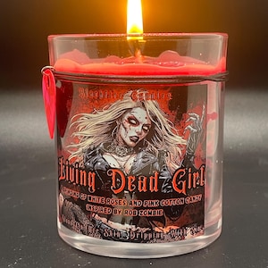 May include: A lit candle in a clear glass jar with a red wax top. The label features a graphic of a woman and the text "Living Dead Girl" with "A Grinding of White Roses and Pink Cotton Candy Inspired by Rob Zombie".