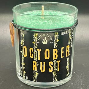 Może przedstawiać: Szklana świeca z zielonym woskiem w środku i drewnianym knotem. Świeca jest oznaczona napisem "October Rust" na czarnym tle z żółtymi literami. Widoczna jest również nazwa marki "Blackfire Candles".