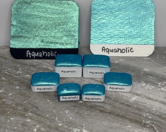 May include: An assortment of watercolor paints in shades of teal and turquoise. The larger paints are rectangular with the word "Aquaholic" written on them. Smaller square paints are also labeled "Aquaholic".