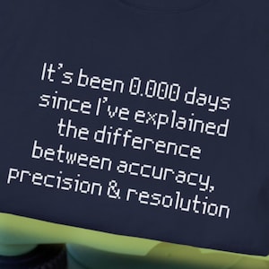 May include: Navy blue t-shirt with white text: "It's been 0.000 days since I've explained the difference between accuracy, precision & resolution." The shirt is displayed on a green and black surveying instrument.
