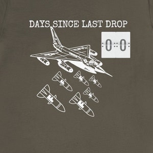 May include: A white graphic on a brown t-shirt shows a jet plane with bombs dropping from it. The text "DAYS SINCE LAST DROP" is above the plane and a digital clock reads "0:00".