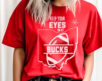 May include: Red t-shirt with a white outline of the state of Ohio and a football with the word "BUCKS" inside. The text "KEEP YOUR EYES ON MY" is above the football.