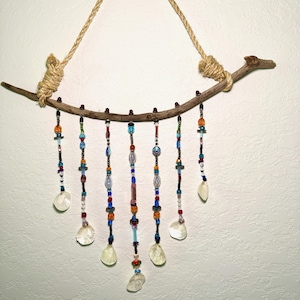 Puede incluir: Un colgante de pared artesanal con una rama de madera natural y una cuerda de yute para colgar. Cadenas de cuentas coloridas de varias formas y tamaños cuelgan de la rama, acentuadas con cristales transparentes en forma de lágrima. Para decoración del hogar.