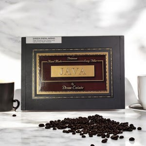May include: A black wooden box with a gold label that reads "Hand Made Premium Long Filler JAVA by Drew Estate". The box is sitting on a white marble surface with coffee beans scattered around it.