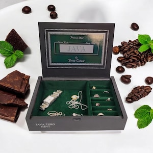 May include: A dark gray wooden box with a green velvet interior. The box has a label that reads "Premium Mint Hand Made - Every Time JAVA by Drew Estate". The box is open and contains a watch, a necklace, and several rings. The box is surrounded by coffee beans and mint leaves.
