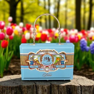 May include: A light blue box-shaped handbag with a decorative gold handle. The handbag features a gold and blue label with the text "My Father Cigars" and "Blue". The background is a blurred garden with colorful flowers.