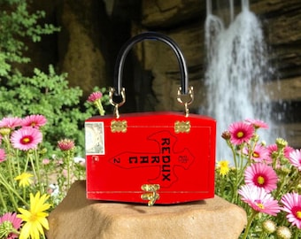 May include: A vibrant red handbag with a black handle and gold-colored hardware. The bag features the word "REDUX" in black lettering and is set against a backdrop of flowers and a waterfall.