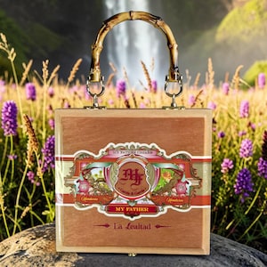 May include: A square wooden box purse featuring a My Father Cigars label on the front with red, gold, and green accents. The box has a curved bamboo handle attached with gold-toned metal hardware and a small metal latch at the bottom edge.