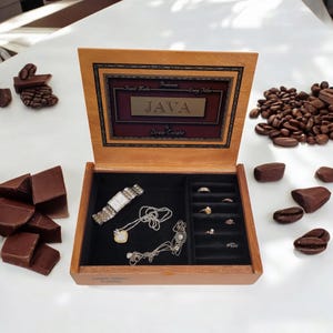 May include: A wooden box with a black velvet interior, containing a watch, two necklaces, and a ring holder with several rings. The box is labeled "JAVA Drew Estate Premium Hand Made Long Filler".