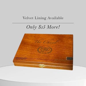 May include: A wooden box with a hinged lid, featuring the text "The Chisel" and an emblem. The box has a warm, brown finish and a gold-colored clasp. The text "Velvet Lining Available" and "Only $15 More!" are above the box.