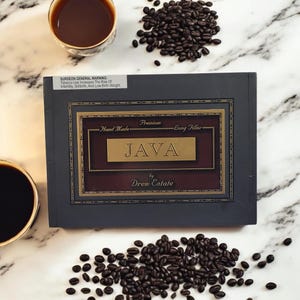 May include: A dark gray box with a gold label that reads "Premium Hand Made Long Filler JAVA by Drew Estate". The box is surrounded by coffee beans on a white marble surface.
