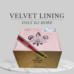 May include: A wooden cigar box with a decorative fleur-de-lis design and the text "Tatuaje La Selección de Cazador Corojo". The box has a red and gold label and the text "25 Cohetes Corojo". The text "VELVET LINING" is above the box.