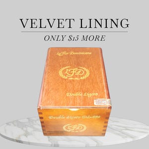 May include: A wooden cigar box with gold lettering and the text "VELVET LINING" and "ONLY $15 MORE". The box has the words "La Flor Dominicana" and "Double Ligero" printed on it. The box is sitting on a white marble surface.