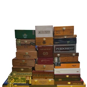 May include: A stack of various wooden cigar boxes in different colors and sizes. The boxes feature printed text and logos, including "BRICK HOUSE", "LIGA PRIVADA", and "PERDOMO". Some boxes have metal clasps.