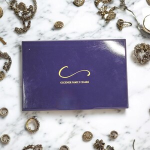 May include: A purple box with a gold logo that says "Ozgener Family Cigars". The logo is a stylized letter "C" with a swirl.