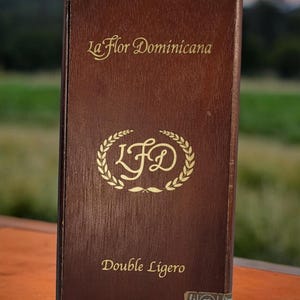 May include: A brown wooden box with gold lettering that reads "La Flor Dominicana" and "Double Ligero". The box has a gold laurel wreath with the letters "LFD" inside.
